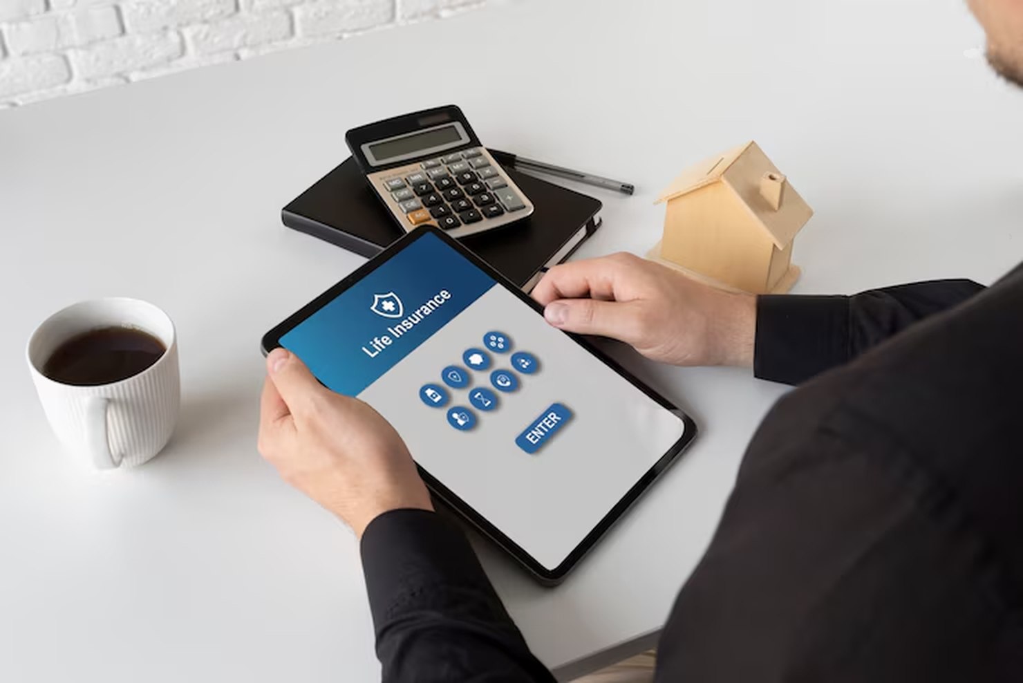 A person holding a tablet with a life insurance interface on the screen, surrounded by a calculator, coffee cup, and miniature house, illustrating how digital tools can help you automate your finances for better planning and peace of mind.