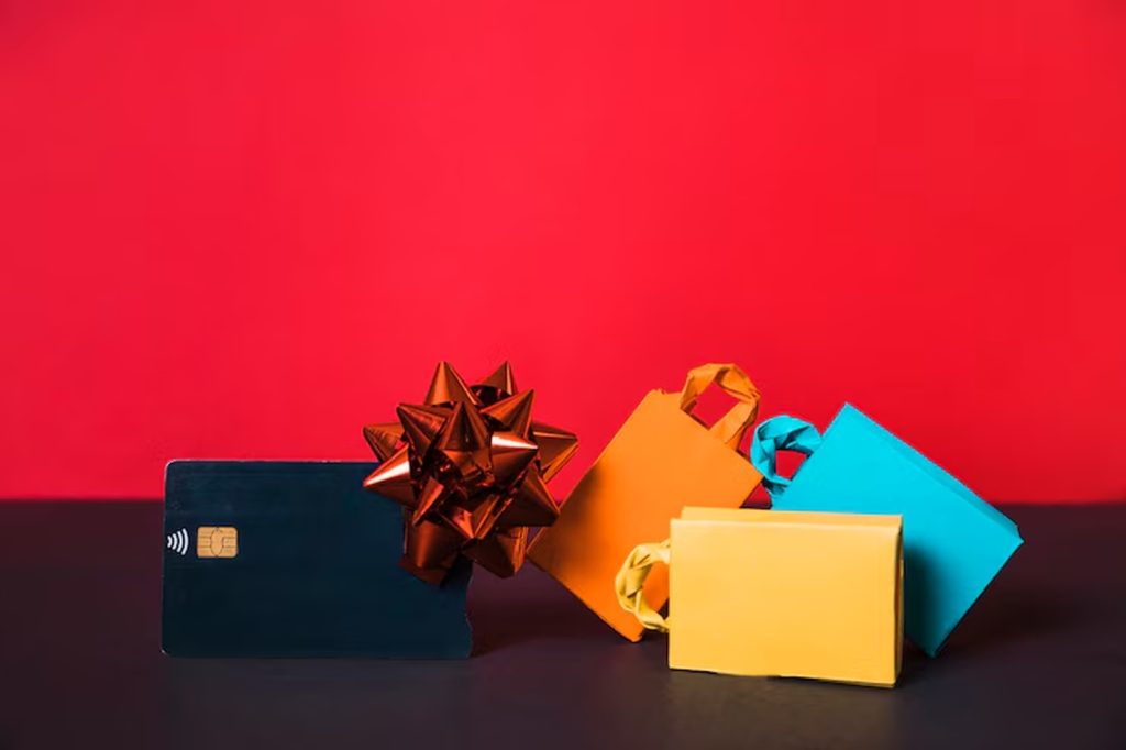 Colorful shopping bags and a gift-wrapped contactless payment card are arranged against a vibrant red background, representing festive spending and the growing popularity of debit cards for everyday purchases.