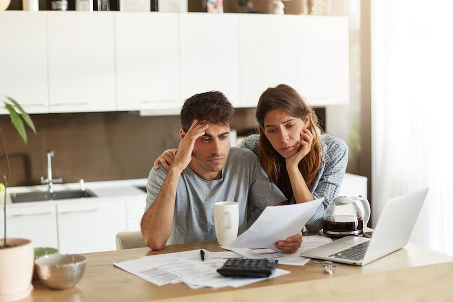 A stressed couple sits at their kitchen table reviewing bills, surrounded by paperwork, a laptop, and a calculator, illustrating the emotional and financial strain that can lead to considering debt consolidation as a solution.