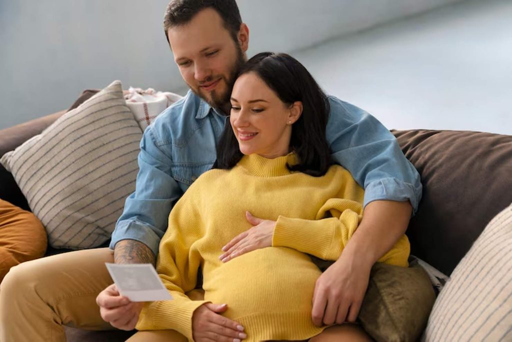 A happy expecting couple sits together on a cozy couch, smiling as they look at an ultrasound photo, symbolizing love, preparation, and future planning related to growing a family and managing their finances.