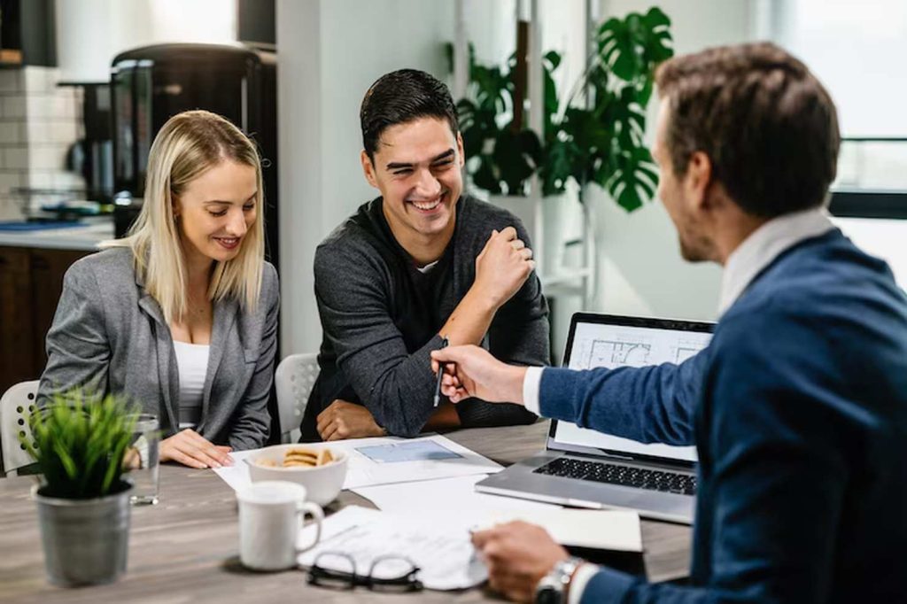 A young couple sits across the table from a professional in a business setting, smiling and discussing documents and plans, symbolizing the supportive role of financial advisors in helping clients make informed decisions about their future.