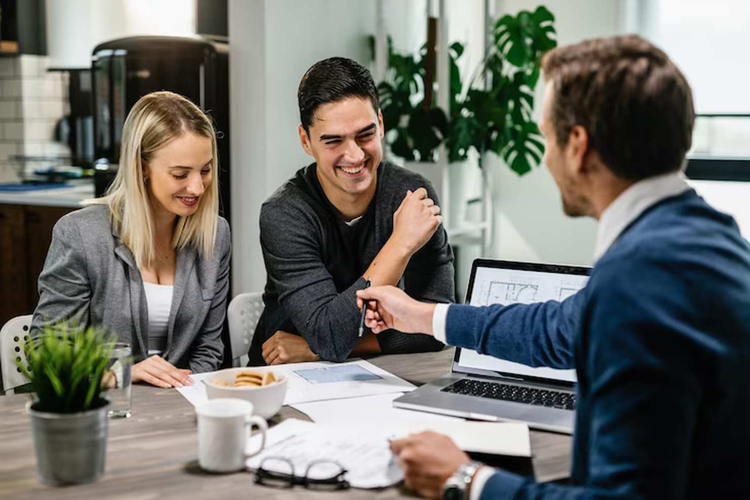 A young couple sits across the table from a professional in a business setting, smiling and discussing documents and plans, symbolizing the supportive role of financial advisors in helping clients make informed decisions about their future.