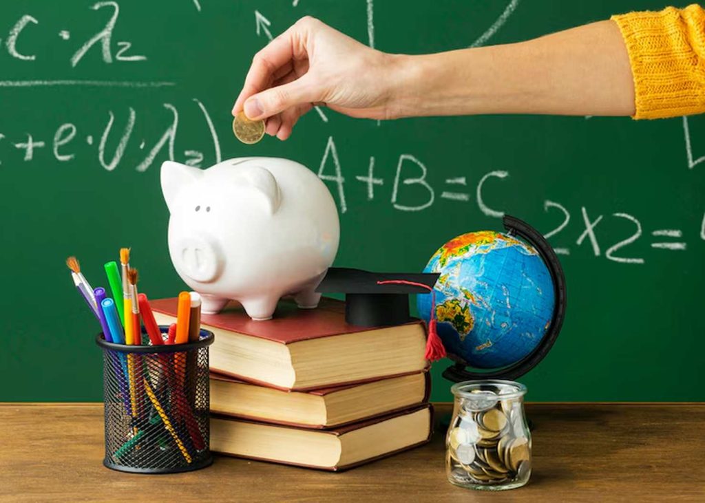 Hand placing a coin into a white piggy bank on top of stacked books, with school supplies, a globe, and a graduation cap nearby, symbolizing the importance of saving and promoting financial education from an early age.