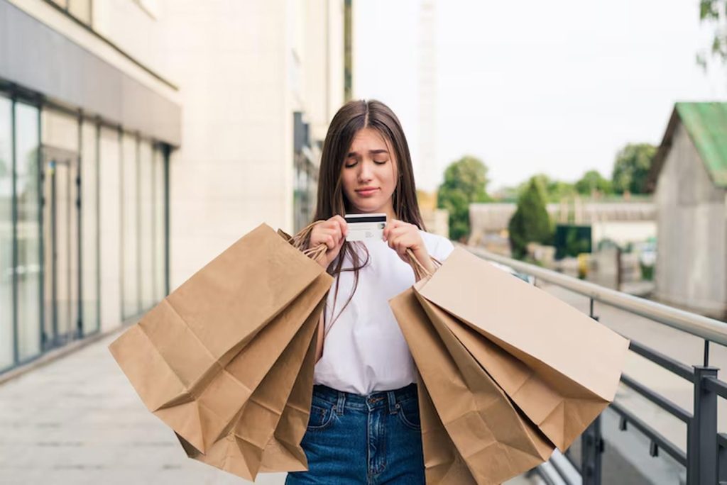 A young woman stands outdoors holding several large shopping bags in both hands while looking down unhappily at her credit card, illustrating the emotional aftermath of impulsive shopping.