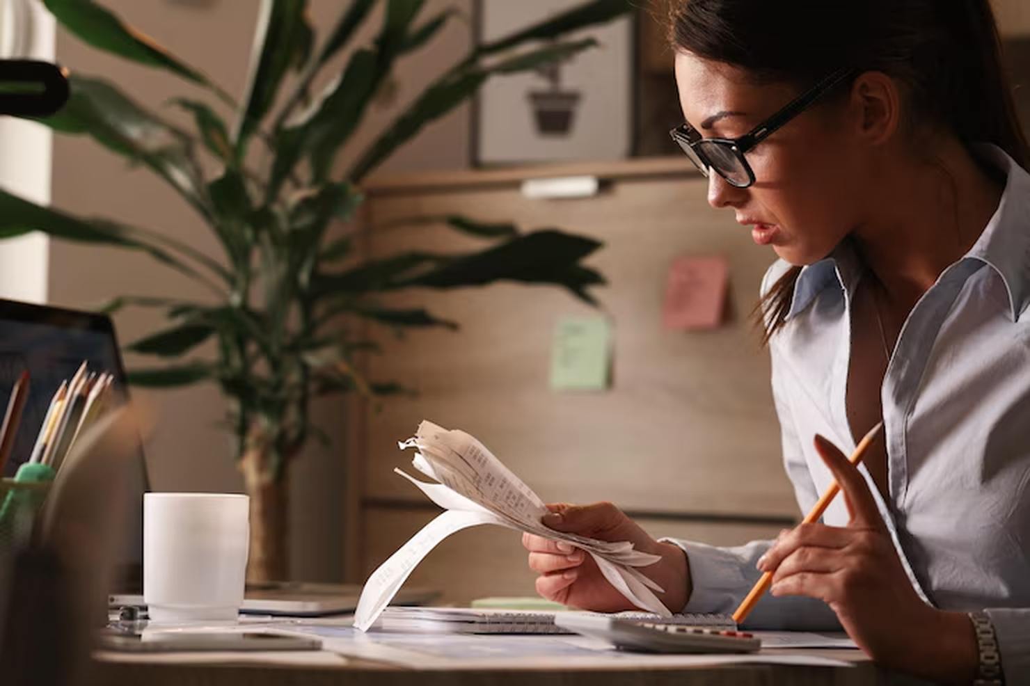 A focused woman sits at a desk reviewing receipts and using a calculator, surrounded by office supplies and documents, representing the effort to plan financially and maintain control over personal expenses.