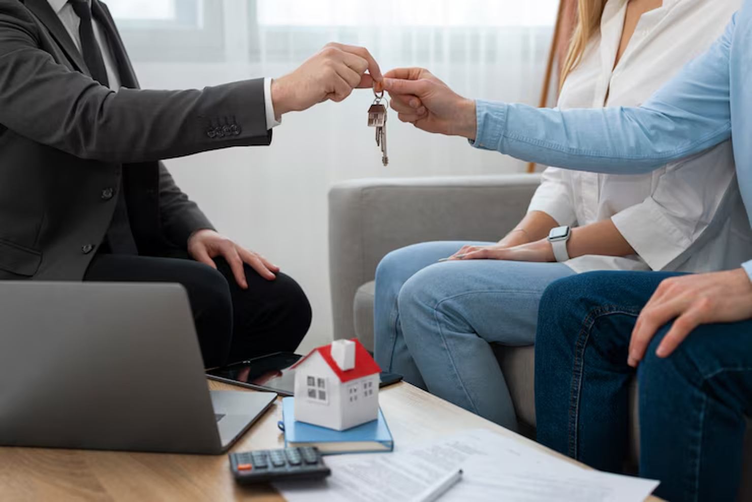 A real estate agent hands over house keys to a couple seated on a couch, symbolizing a successful property buying process, with documents, a laptop, and a small model house on the table nearby.