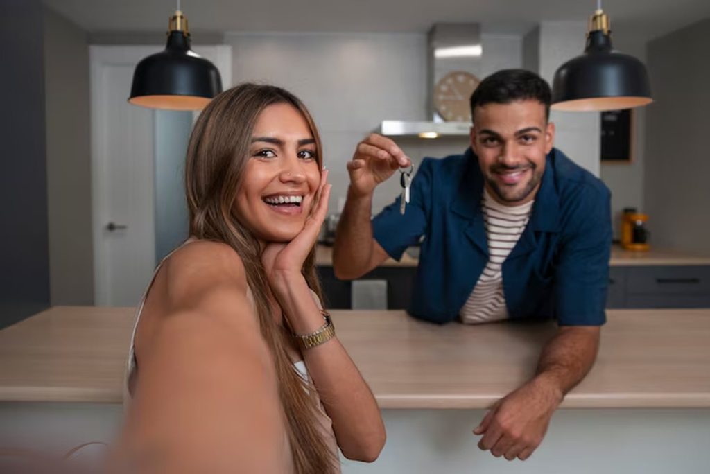 A happy couple stands in their new kitchen, smiling and holding keys while taking a selfie, celebrating their decision to rent or buy a house and start a new chapter together.