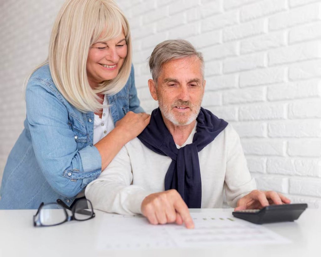 Smiling senior couple reviewing financial documents together at a table, with the man using a calculator and the woman leaning over his shoulder, illustrating thoughtful planning for retirement.