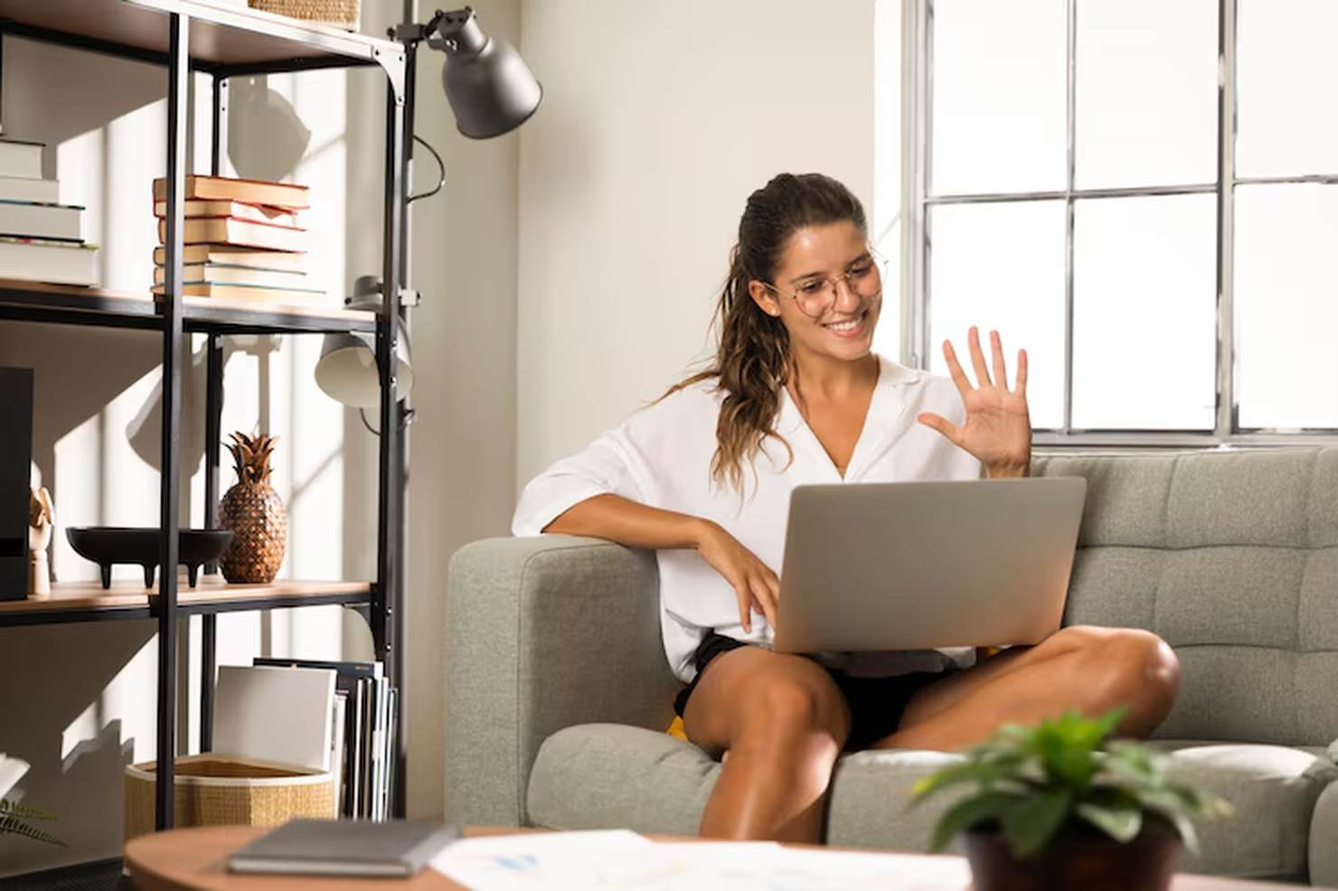 A smiling young woman sits comfortably on a couch, waving at her laptop during a virtual meeting, representing the modern remote lifestyle often associated with flexible side hustles.
