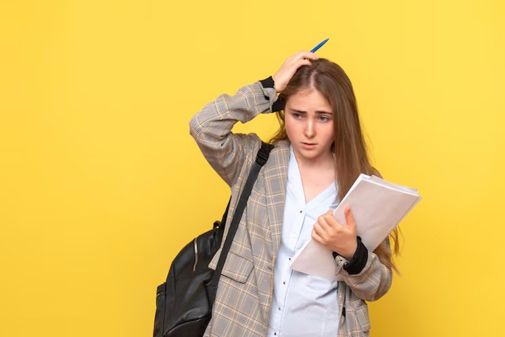 Worried young student holding documents and scratching her head, with a backpack over one shoulder, standing against a yellow background, symbolizing the stress and confusion often associated with student debt.