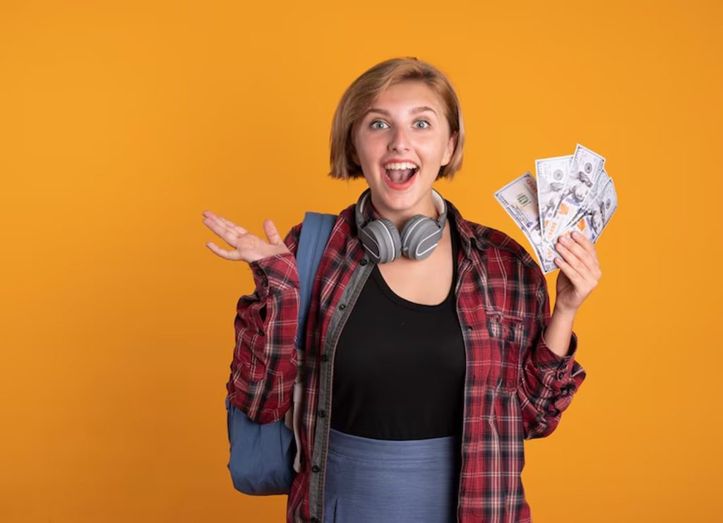Excited young student holding a fan of dollar bills while wearing a backpack and headphones, standing against an orange background, symbolizing the positive impact of receiving or managing student loans.