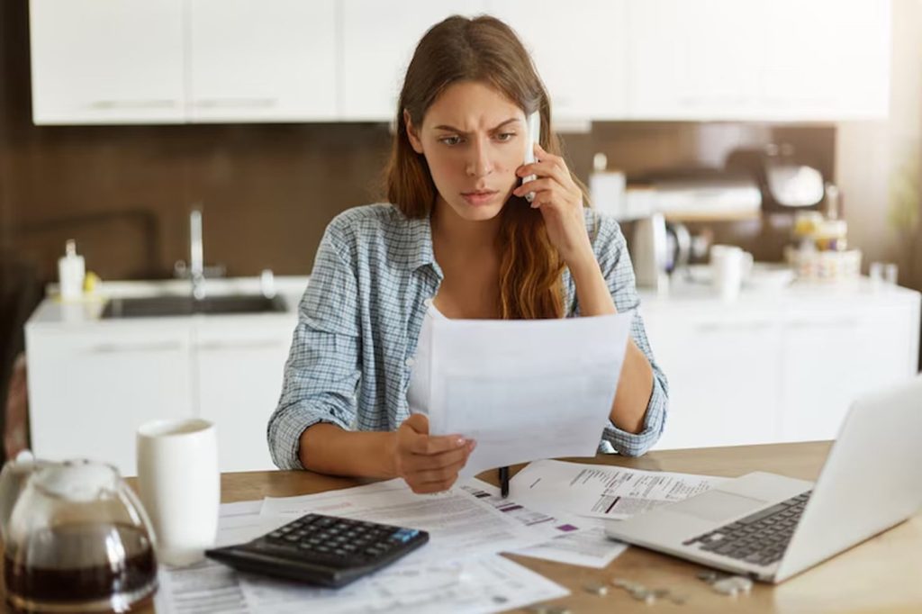 A concerned young woman talks on the phone while reviewing bills at her kitchen table, surrounded by documents, a calculator, and a laptop, reflecting the stress of dealing with unexpected expenses.