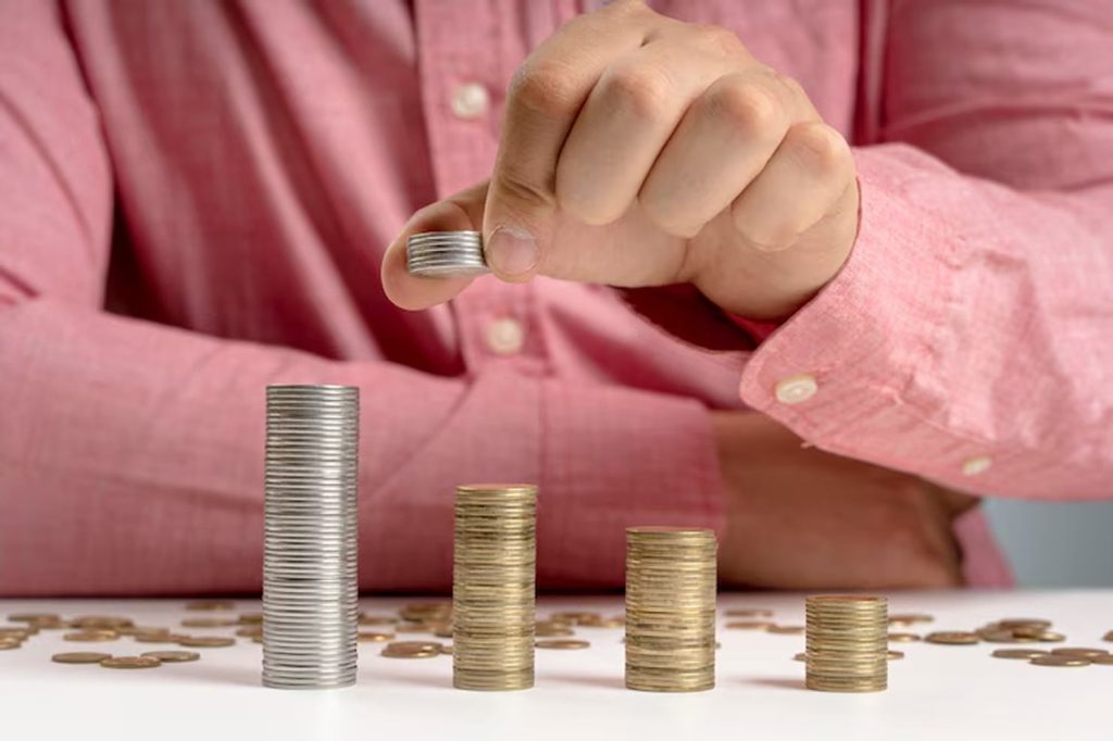A person in a pink shirt stacks coins of different sizes on a white table, representing the challenge of managing savings and budgeting with variable income.