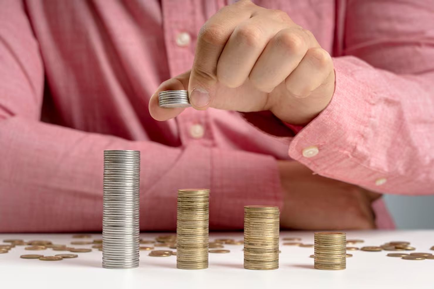 A person in a pink shirt stacks coins of different sizes on a white table, representing the challenge of managing savings and budgeting with variable income.
